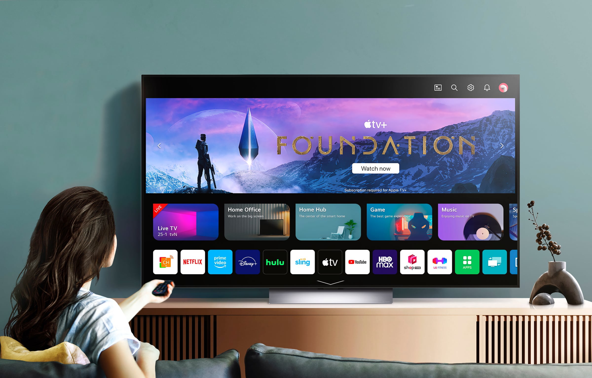 Woman in her living room watching the webOS home screen on an LG Smart TV, featuring Apple TV+ Foundation, shortcuts to Live TV, Home Hub, Game and Music, and apps like Netflix, Prime Video, Disney+ and HBO Max